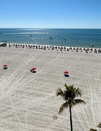 Aerial view of Fort Myers Beach with white sand, rows of beach umbrellas, palm tree foreground, and calm Gulf of Mexico waters.
