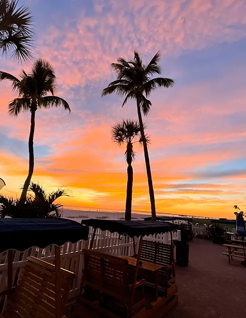 Vibrant sunset over Fort Myers Beach with palm trees, pastel skies, and outdoor seating near the shoreline.